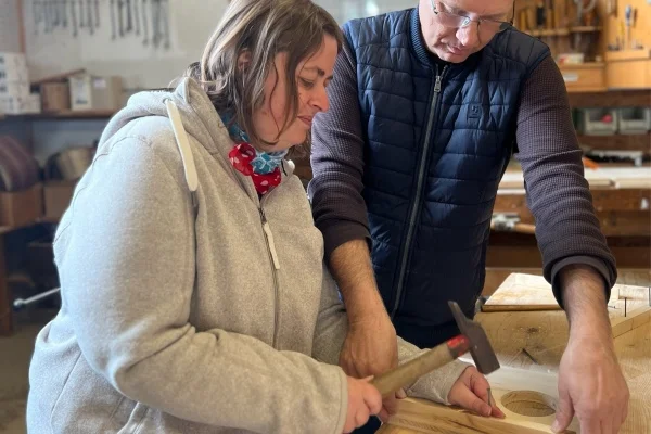 Das Bild zeigt eine Frau, die mit Hilfe eines Mannes einen Nagel in ein Brett schlägt