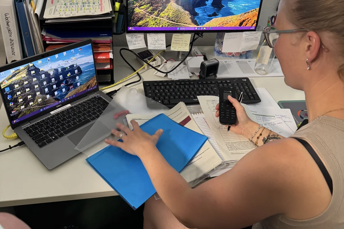 Familienfreundliche Arbeitsgestaltung Eine Frau sitzt an einem vollen Schreibtisch, vor einem Laptop und hält ein Telefon in der Hand.