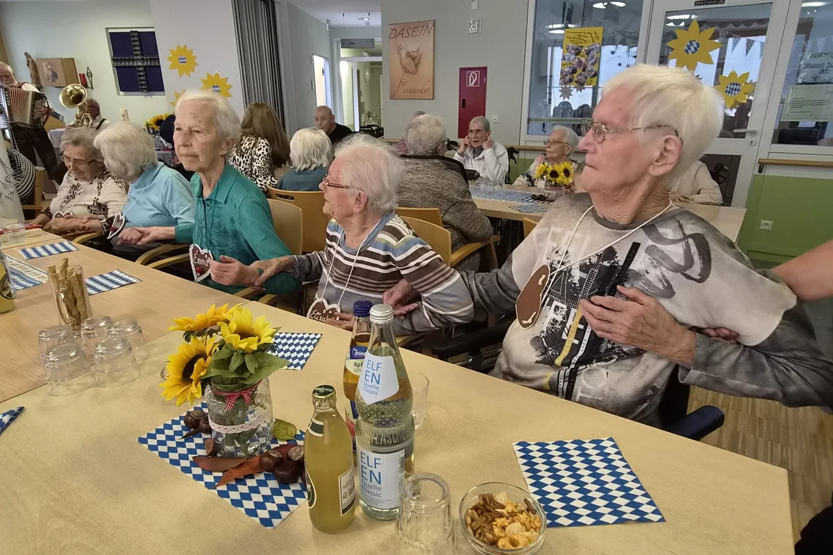 Gruppe älterer Menschen in einem Seniorenheim sitzt an einem festlich gedeckten Tisch mit Sonnenblumen, blauen Servietten, Getränken und Snacks; zwei Frauen halten einander die Hände und blicken aufmerksam.
