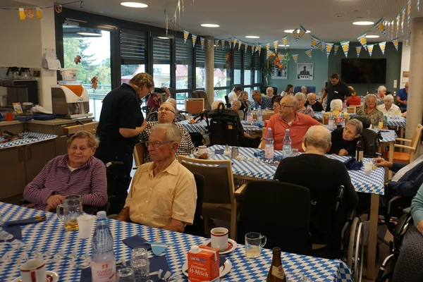 Gruppe älterer Menschen in festlich dekoriertem Gemeinschaftsraum, mehrere Tische mit blau-weiß karierten Tischdecken, Getränke und Gespräche.