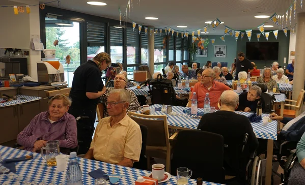 Gruppe älterer Menschen in festlich dekoriertem Gemeinschaftsraum, mehrere Tische mit blau-weiß karierten Tischdecken, Getränke und Gespräche.