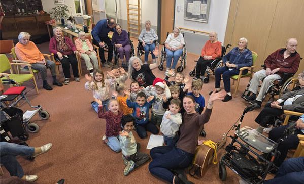 Eine Gruppe Kindergartenkinder sitzt in der Mitte eines Stuhlkreises mit vielen Bewohnerinnen und Bewohnern des Pflegeheims.