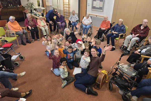 Besuch aus dem Kindergarten Eine Gruppe Kindergartenkinder sitzt in der Mitte eines Stuhlkreises mit vielen Bewohnerinnen und Bewohnern des Pflegeheims.