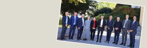 Mehrere Personen in formeller Kleidung stehen nebeneinander auf einem gepflasterten Weg vor Bäumen und Sträuchern, lächelnd, Gruppenporträt bei Tageslicht.