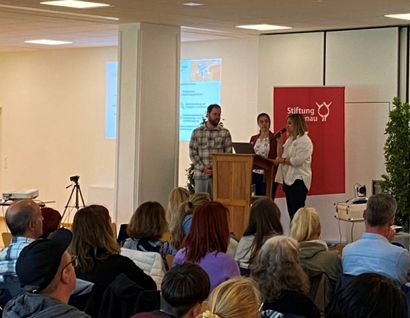 Das Bild zeigt ein Podium mit Vortragenden und Beamer vor Publikum