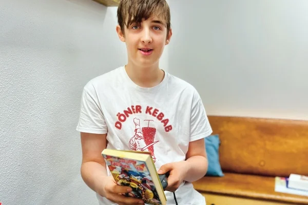 Den Leihverkehr verwalten die Schülerinnen und Schüler selbstständig – auch dank digitaler Technik Das Bild zeigt einen Jungen der ein T-Shirt anhat auf dem in rot Döner Kebap steht und er hält ein Buch in der Hand.