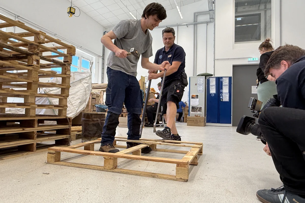 Paletten Drei Männer in einer Werkhalle reparieren oder montieren Holzpaletten; einer schlägt mit einem Hammer auf den Palettenrahmen, ein weiterer hält die Palette, neben ihnen kniet ein Fotograf und macht Fotos.