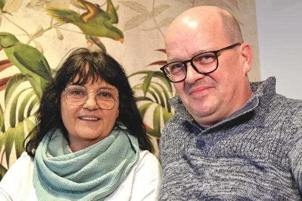 das Bild zeigt eine Frau und einen Mann mit Brille die gemeinsam vor der Kamera stehen und in die Kamera lächeln