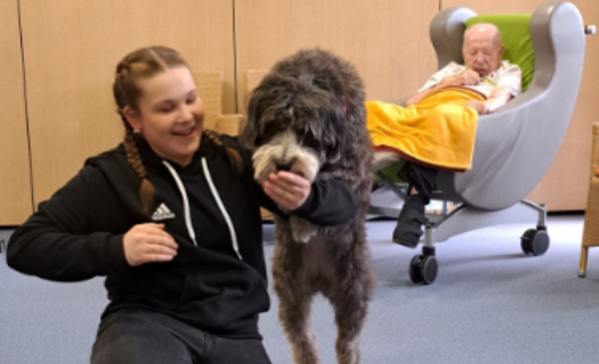 Das Bild zeigt im Vordergrund eine junge Frau mit Hund, im Hintergrund einen Mann in einem Sessel