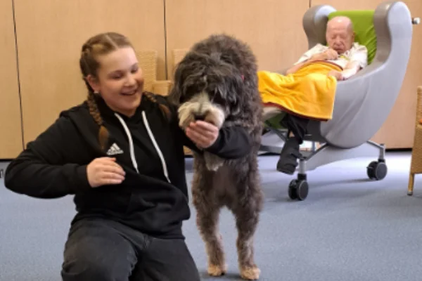 Das Bild zeigt im Vordergrund eine junge Frau mit Hund, im Hintergrund einen Mann in einem Sessel