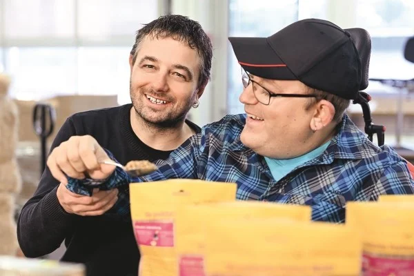 Was bedeutet Arbeit für den Menschen? Das Bild zeigt einen Menschen mit Behinderung bei der Arbeit, assistiert von einem Betreuer