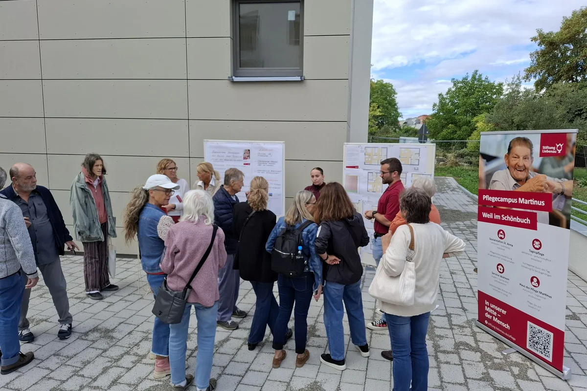 Tag der offenen Baustelle Gruppe von rund zwölf Erwachsenen steht vor einer Gebäudewand im Freien und sieht sich Informationsplakate sowie ein rotes Aufsteller-Banner zu Pflegeangeboten an.