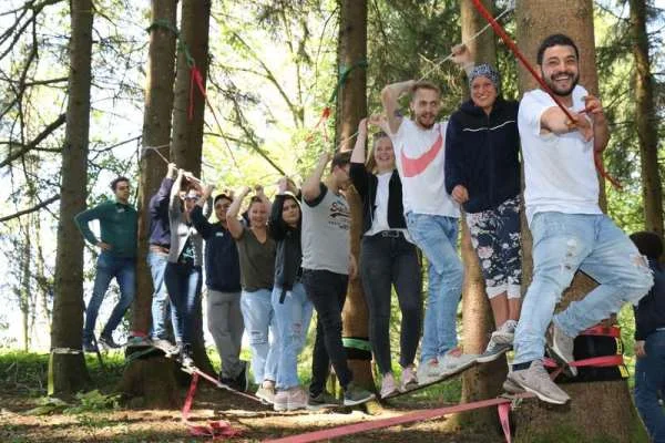 Das Bild zeigt eine Gruppe von Menschen, die auf einem Seil stehen das zwischen zwei Bäumen gespannt ist