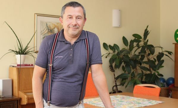 Das Bild zeigt einen Mann in Hosenträgern der an einem Tisch lehnt und in die Kamera lächelt