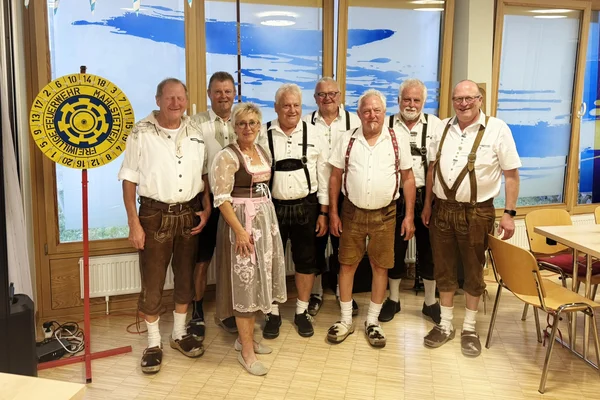 Neun Personen, überwiegend Männer in Lederhosen und eine Frau im Dirndl, stehen lächelnd in einer Reihe in einem Raum mit blau-weißer Oktoberfest-Dekoration.