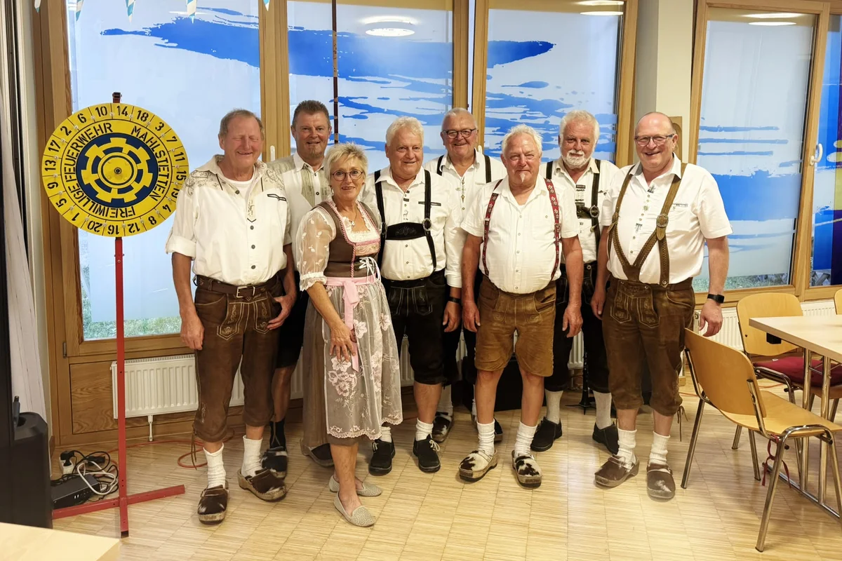 Neun Personen, überwiegend Männer in Lederhosen und eine Frau im Dirndl, stehen lächelnd in einer Reihe in einem Raum mit blau-weißer Oktoberfest-Dekoration.