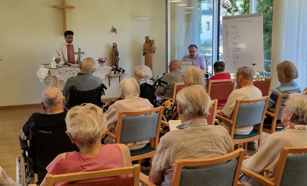 Patrozinium im Haus der Pflege St. Dominikus in Bad Grönenbach Mehrere Personen sitzen auf Stühlen und schauen zu einem Mann in einem Gewand der zu ihnen redet.