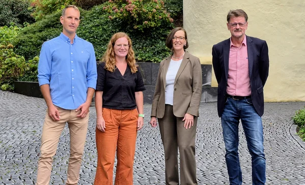 Vier Erwachsene — zwei Männer und zwei Frauen — stehen nebeneinander auf einem Kopfsteinpflasterweg vor dem schloss Liebenau, in lässig-formeller Kleidung in die Kamera blickend.