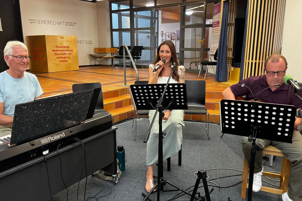 Absolventenfeier Das Bild zeigt eine singende Frau, während rechts ein Mann Gitarre spielt und links ein Mann am Klavier sitzt.