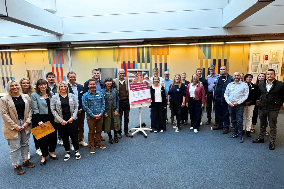 Zukunftstag Gruppe von etwa 24 Personen, formell bis lässig gekleidet, steht in einem hellen Innenraum im Halbkreis um ein auf einem Ständer platziertes Plakat.