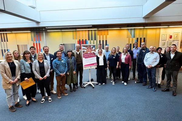 Gruppe von etwa 24 Personen, formell bis lässig gekleidet, steht in einem hellen Innenraum im Halbkreis um ein auf einem Ständer platziertes Plakat.