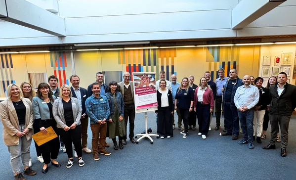 Gruppe von etwa 24 Personen, formell bis lässig gekleidet, steht in einem hellen Innenraum im Halbkreis um ein auf einem Ständer platziertes Plakat.