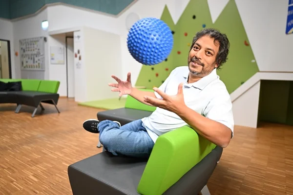 Das Bild zeigt einen Mann der auf einer Bank sitzt und einen blauen Ball in die Luft wirft.