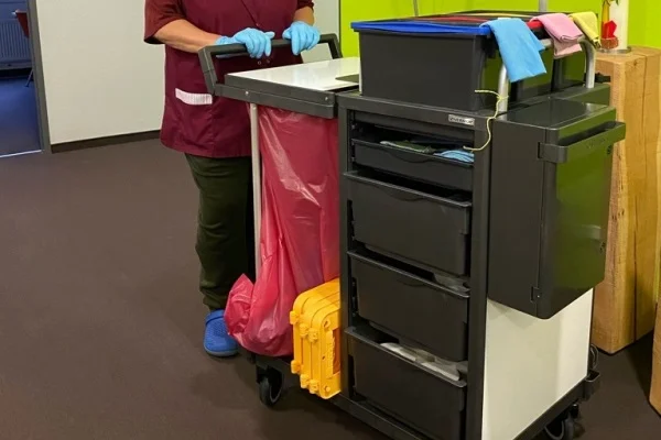 Das Bild zeigt eine Person mit Handschuhen, die gerade einen Putzwaagen vor sich herschiebt