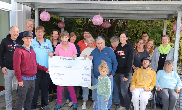 Gruppe von etwa 20 Menschen, darunter Erwachsene und Kinder, steht auf einer Terrasse und hält gemeinsam einen großen symbolischen Spendenscheck für eine Stiftung, alle lächelnd.