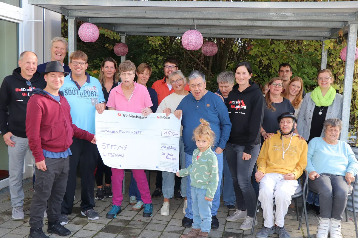 Spende Hypovereinsbank Scheckübergabe Gruppe von etwa 20 Menschen, darunter Erwachsene und Kinder, steht auf einer Terrasse und hält gemeinsam einen großen symbolischen Spendenscheck für eine Stiftung, alle lächelnd.