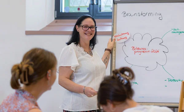 Eine Frau zeigt an einer Tafel etwas und zwei andere Personen hören ihr zu.