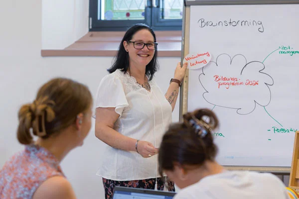 Eine Frau zeigt an einer Tafel etwas und zwei andere Personen hören ihr zu.