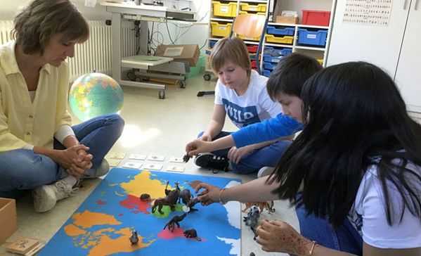 das bild zeigt die montessori-lehrerin mit einer einer gruppe an schülern am boden auf einem teppich lernend