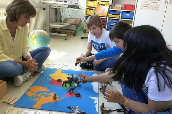 das bild zeigt die montessori-lehrerin mit einer einer gruppe an schülern am boden auf einem teppich lernend