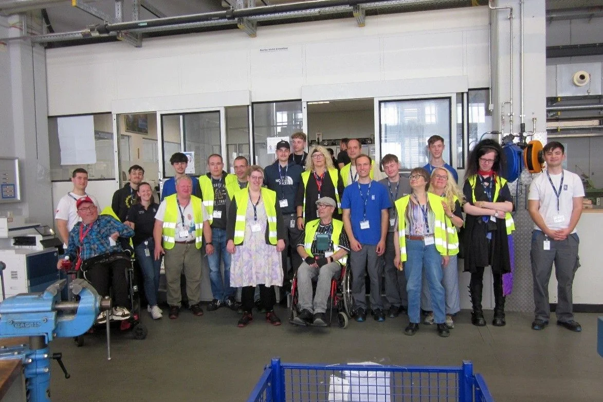 Tandem-Projekt von der Stiftung Liebenau und Rolls Royce Power Systems Gruppenbild verschiedener Personen in Warnwesten in einem Gebäude.