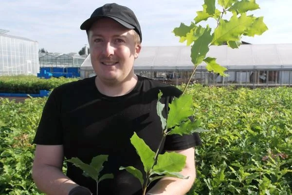 Das Bild zeigt einen jungen Mann mit einer Baumpflanze vor einem Feld
