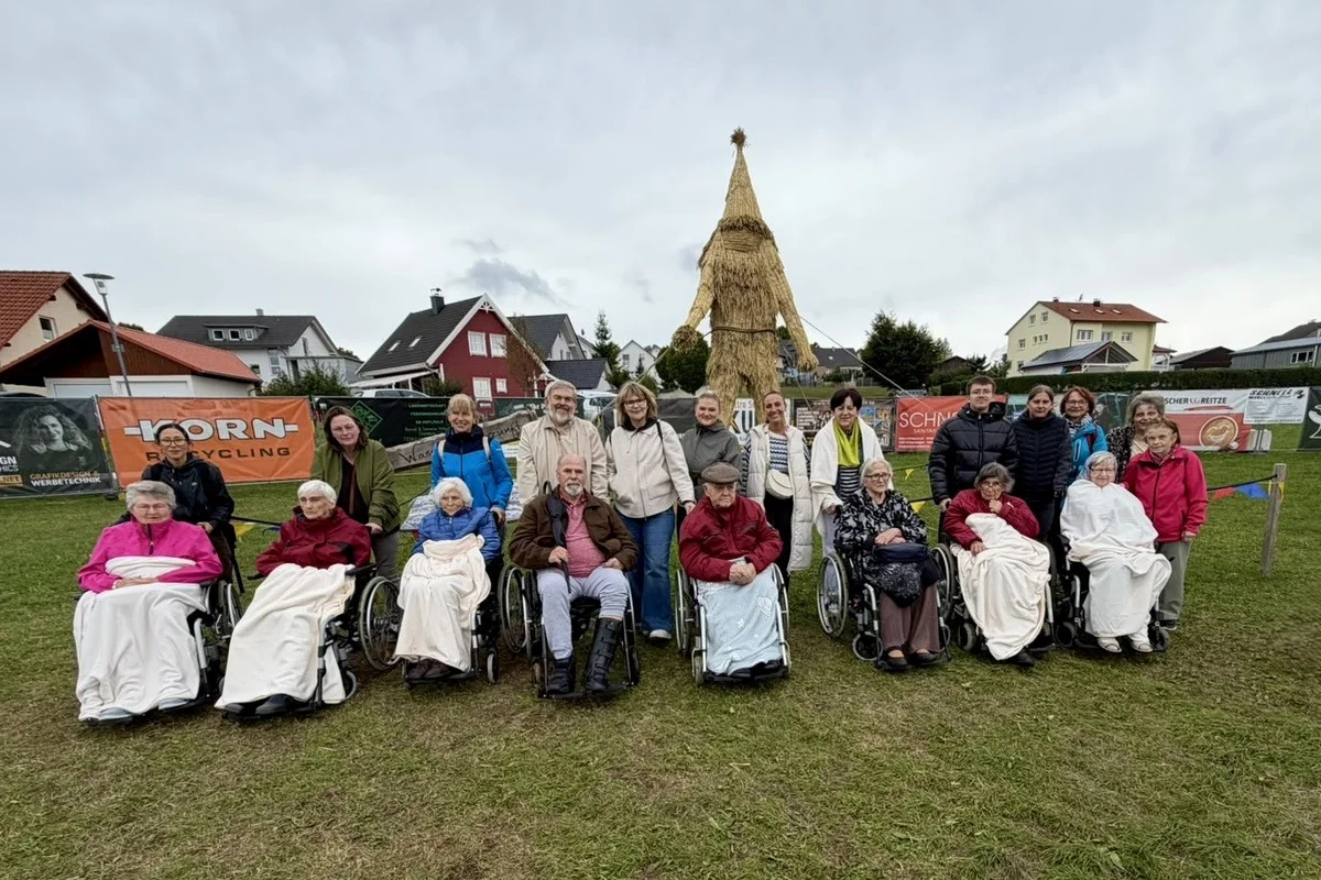 Bewohnerausflug Strohpark Gruppenfoto mehrerer Menschen, einige in Rollstühlen, sitzend und stehend auf einer Wiese vor einer großen Strohpuppe; Wohnhäuser im Hintergrund und bewölkter Himmel.
