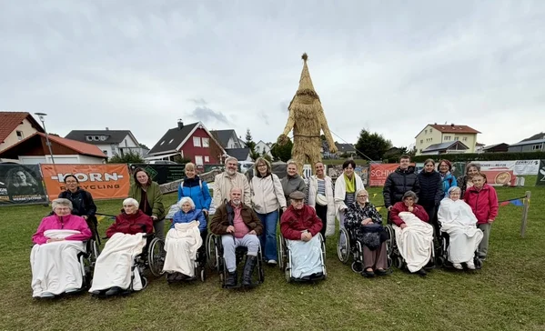 Bewohnerausflug Strohpark Gruppenfoto mehrerer Menschen, einige in Rollstühlen, sitzend und stehend auf einer Wiese vor einer großen Strohpuppe; Wohnhäuser im Hintergrund und bewölkter Himmel.