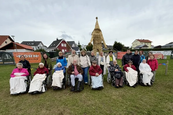 Gruppenfoto mehrerer Menschen, einige in Rollstühlen, sitzend und stehend auf einer Wiese vor einer großen Strohpuppe; Wohnhäuser im Hintergrund und bewölkter Himmel.