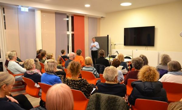 Das Bild zeigt einen Raum in dem viele Personen sitzen auf orangenen Stühlen und einem Vortrag zuhören