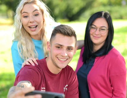 Das Bild zeigt einen Mann, der zwischen zwei Frauen steht. Zu dritt lächeln sie in die Kamera und sehen glücklich aus.