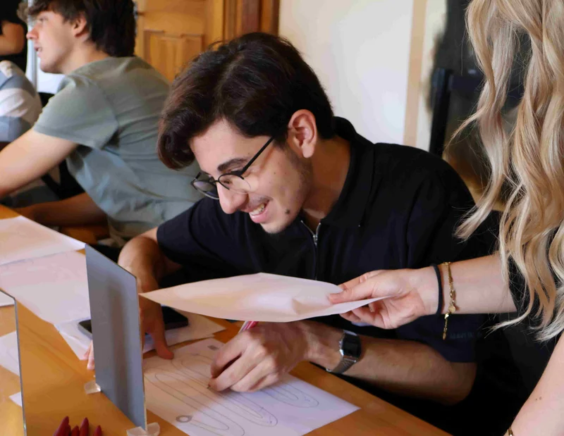 Einführungstag 2026 Ein Mann schreibt auf einem Papier, indem er spiegelverkehrt denken muss, da ein Spiegel vor ihm steht. Eine Frau reicht ihm ein weiteres Papier.