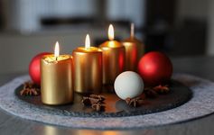 Das Bild zeigt vier goldene Kerzen die auf einem grauen Untersetzer stehen und brennen. Neben ihnen liegen Weihnachtskugeln in weiß und rot 