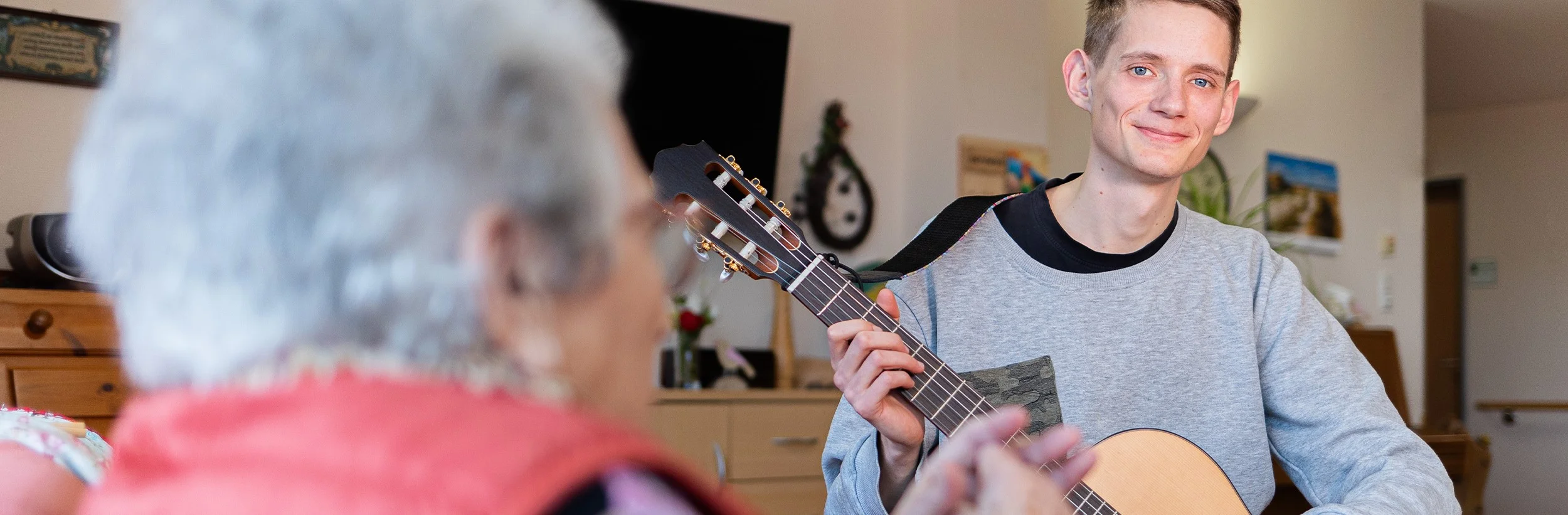 Das Bild zeigt einen Mann der Gitarre für eine ältere Dame spielt. Der Mann lacht in die Kamera