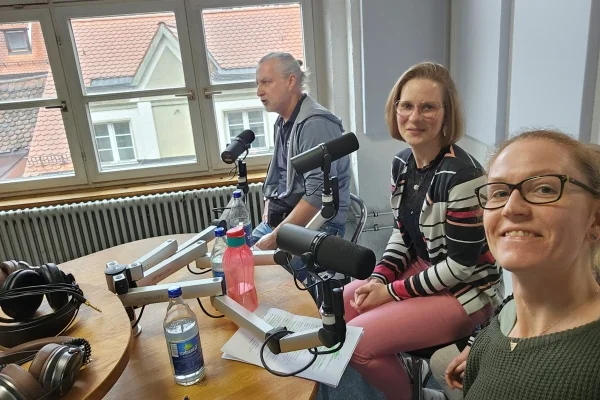 Das Bild zeigt drei Personen vor einem Mikrofon