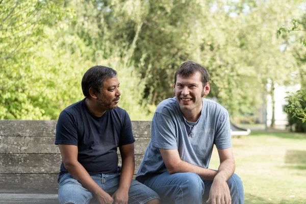 Das Bild zeigt zwei Männer die auf einer Parkbank auf einer Wiese sitzen