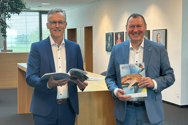 Zwei Männer halten eine Broschüre in der Hand und lächeln in die Kamera.