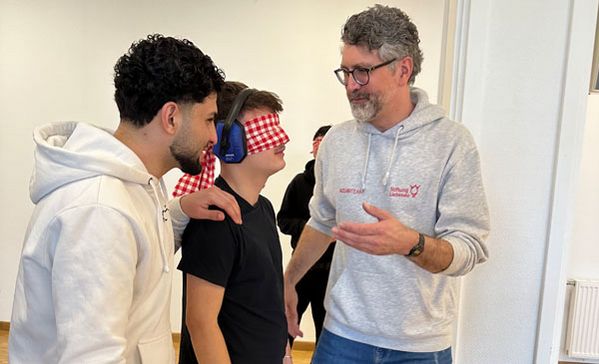 Besuch von Schülerinnen und Schülern Zwei Personen führen einen Jugendlichen mit Gehörschutz und Augenbinde durch einen Raum.