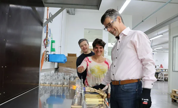 Ein Mann mit Schutzbrille arbeitet und zwei Personen stehen im Hintergrund.