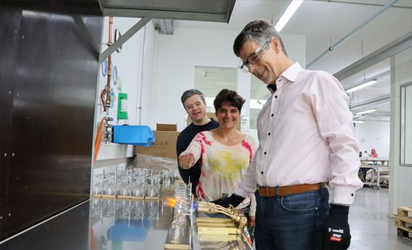 Ein Mann mit Schutzbrille arbeitet und zwei Personen stehen im Hintergrund.